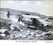 A black and white image a tree-less rocky terrain. A early 20th century ambulance is in the middle of the image, on a road with on an incline, facing to the right, down the incline. There is one person behind the ambulance pushing it. there is another person standing a few metres behind.