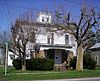 A white wooden house with a porch, situated against a blue sky. Two leafless trees stand between the house and the camera.