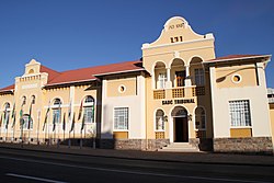 A yellow and white neo-classicist building in bright sunlight. The roof of corrugated iron sheets is painted dark red, on the main gable the inscriptions read "A.D.1913" and "SADC Tribunal". Seven flagpoles with national flags of countries from the Southern African Development Community are mounted onto the pavement in front of the building.