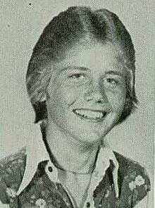 Monochrome bust photo of a blond-haired white teen wearing an open-collared shirt; he is smiling and looking into the camera.