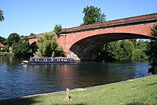 A wide tree-lined river on the left has a green bank in the right-foreground and is crossed by a low two brick-arched bridge. A tree in the middle of the river obscures part of the bridge.