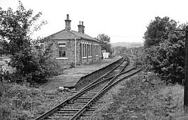 A black and white image of a single storey station building, platform and two lines that merge near to the camera into one line