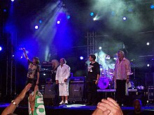 Scène de concert avec quatre hommes et une femme sur la scène devant une foule.
