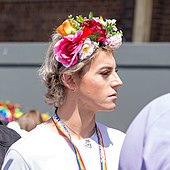 Man seen in profile wearing makeup, with flowers arranged on a headband high on his head; a rainbow-lolored lanyard around his neck reads "Suffolk Pride"
