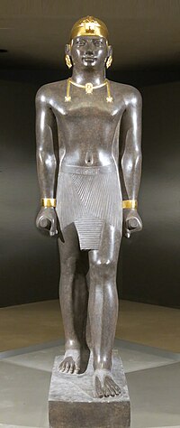 Statue of Taharqa. His name appears on the center of his belt: 𓇿𓉔𓃭𓈎 (tꜣ-h-rw-q, "Taharqa"). The statue is 2.7 meters tall. Taharqa has a striding pose, the arms held tight, and holds the mekes staff. He wears a shendyt or pleated kilt and on his head is a double-uraeus skullcap, possibly signifying his rule over Nubia and Egypt.[1] (Louvre Museum, color reconstruction of the jewelry through pigment analysis).[2]