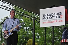 McCotter on stage with a guitar beside a campaign poster