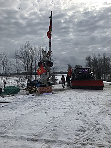Mohawk protesters stand by a snowplow covered in Iroquois and Mohawk Warrior flags near a level crossing at Wyman Rd with firewood and a wooden sign that reads "#RCMP get out".