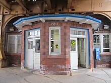 A brick station house, at street level, with an angled bay window. The windows have white trimming, while the doors and house cornice have blue trimmings.