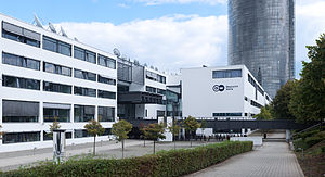 A white building complex with the logo of Deutsche Welle. The background shows a skyscraper made out of glass.