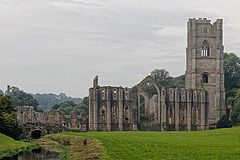 A photograph of a ruined abbey; a river passes by in the lower left hand of the picture, overhung with dark trees. A ruined abbey building in stone makes up the midground of the right side of the photograph.