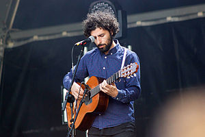 José González wearing a dark blue shirt, standing onstage in front of a microphone, playing an acoustic guitar