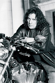 black-and-white image of Pappo wearing jeans and a leather jacket, sitting on a Harley Davidson motorcycle