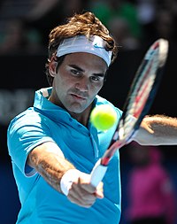 A brown-haired man in a light blue polo shirt and both white wristband and bandanna with a light blue nike logo, who is hitting a backhand with the tennis ball in the foreground