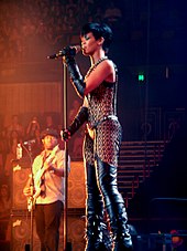 A young woman with black short hair is singing into a microphone on a stand while wearing a leather outfit