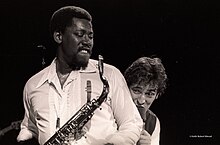 A black-and-white photograph of a man holding a saxophone with another man peering from behind him, singing into a microphone.