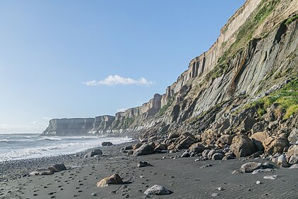 Sur une plage de sable gris ; on aperçoit la mer ; de gigantesques falaises dessinent une baie ; quelques blocs tombés sur la plage.