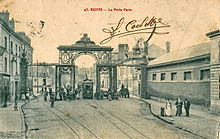 Photographie d'une carte postale en noir et blanc représentant une largue rue barrée d'un porche en ferronnerie.