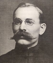 Black and white photo of a dark-haired middle-aged man wearing rounded spectacles and a large bushy moustache