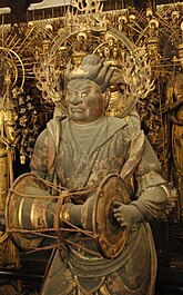 Three-quarter view of a statue carrying a handdrum in front. A halo is visible around his head.
