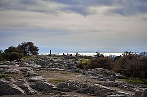 Photo d'un plateau rocheux avec de la végétation et des gens au fond. Au second plan la mer, les montagnes et le ciel.