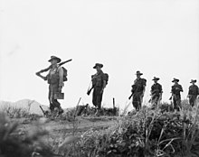 Six evenly spaced Caucasian soldiers wearing slouch hats with their rifles slung march along a dirt road in a straight line from right to left, carrying packs. The camera is at ankle height amidst the grass, which is in the foreground, while the men are silhouetted against the skyline.