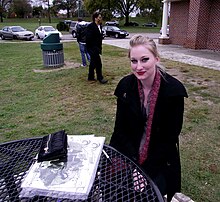 A photo of Abigail Larson. She is a white woman with blonde hair which is pinned up with a clip. She is wearing heavy black eyeliner, a black coat, and a purple scarf. She is sitting outdoors in a park at a table with a stack of posters in front of her. She is looking at the camera and smiling slightly.