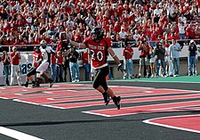 Texas Tech Red Raider football player stands with the a football in the endzone in the foreground with fans in the stands in the background cheering.