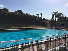 A view of a diving pool filled with water, and a railway embankment behind.