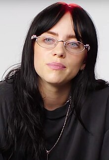 Image of Billie Eilish with long black hair and red roots, wearing silver glasses and black clothes