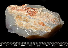 A rock set on a black background with a scale in millimetres at the bottom edge. It shows a mixture of light brown rock with translucent white rock on the underside.