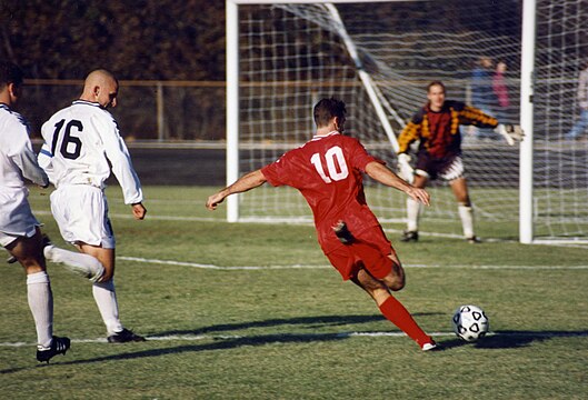 The attacking player (No. 10) attempts to kick the ball beyond the opposing team's goalkeeper, between the goalposts, and beneath the crossbar to score a goal. (Current image)