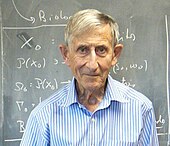 Photograph of Freeman Dyson in front of a blackboard covered in equations