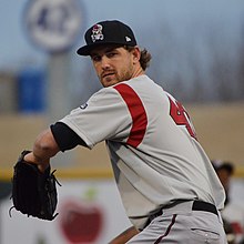 A man wearing a gray baseball jersey with a red stripe on the under arm and red numbers on the back and a black cap and baseball glove prepares to pitch a ball