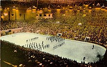 Far view of an ice hockey rink, with ice hockey players and a band standing at its centre. Spectators seated around the rink watch.