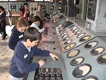 Children fiddling with knobs on a long flat grey metal surface with large dials facing them