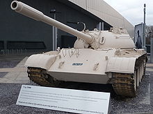 A tank, painted a light desert colour, facing the camera with a large metal-roofed building behind.