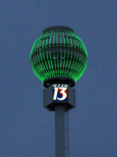 A large steel sphere with neon tubes lit in green, mounted high on a pole bearing the WZZM logo, at night