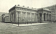 An Egyptian-Revival-style building on a street corner; there are doric columns on the façade to the right of the image. There are men on the street, and work is being done using cranes, on an adjacent building out of frame to the right.