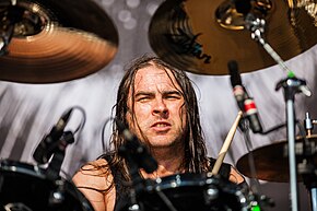 Ryan, aged 28, plays drums while partially looking at the camera. He has a tank top, with all arms hidden behind the drums.