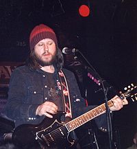 A man playing a guitar and singing on stage. He is wearing a denim jacket and woolen cap