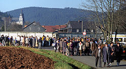 Un grand nombre de personnes de tous ages se trouvent le long d'une route en face d'un mur de béton derrière lequel se trouvent des maisons et une église.