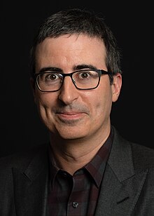Photo of Oliver standing against a black background, wearing glasses and a dark suit jacket.