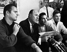 Four men sit on a couch; three look on as the fourth speaks to a group of reporters off camera