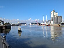 Photo from opposite side of the water to the main bascule span, towed by two boats. It sits on the left side of the harbour by a grain silo building, close to the existing construction of the bridge.