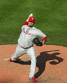 A man wearing a gray baseball uniform with red trim and a red baseball cap throws a baseball with his right hand from a pitcher's mound