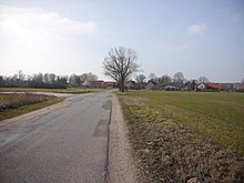 A road through open fields passes a line of trees and enters a village