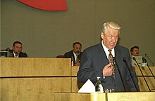 An older white male in a dark suit speaks at a wide wooden podium.