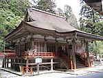 Three-quarter view of a proportionally tall wooden building with a veranda with red hand rail and a canopy covering the steps that lead to the central entrance. The roof appears to be a hip-and-gable roof.