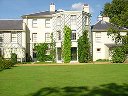 Stucco rendered building in a plain Georgian style with hipped slate roofs and overhanging eaves. The central block is three storeys, and there are two-storey extensions on each side. Lattices are fixed to walls between windows to support climbing plants. A wide lawn forms the foreground, and tall trees appear behind the block to the right.