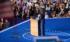 A man, wearing a suite, standing behind two transparent, glass teleprompters.